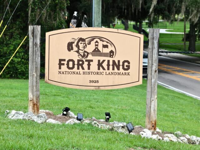 A beige sign held up on either side with wood pillars and lights below reads, "FORT KING NATIONAL HISTORIC LANDMARK 3925." On the sign is a drawing of a fort and s Seminole. In the background is a road with a car and trees.