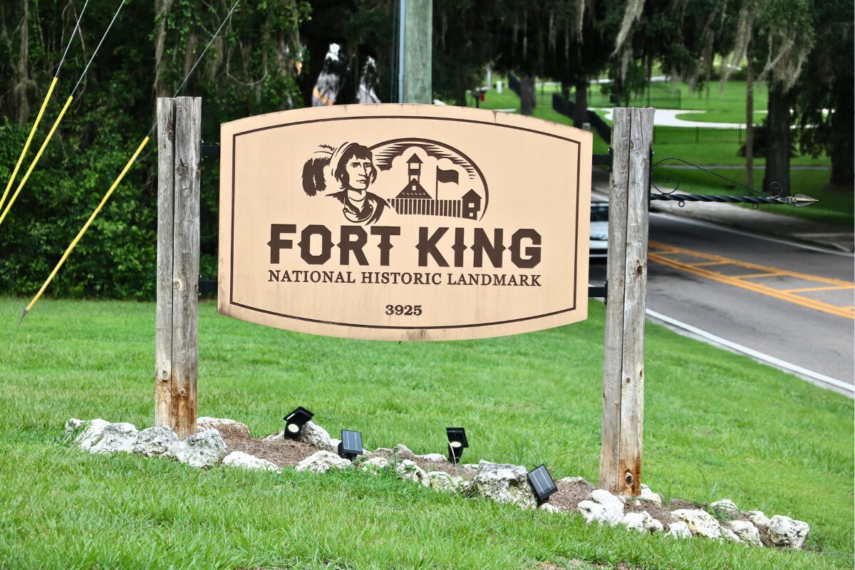 A beige sign held up on either side with wood pillars and lights below reads, "FORT KING NATIONAL HISTORIC LANDMARK 3925." On the sign is a drawing of a fort and s Seminole. In the background is a road with a car and trees.