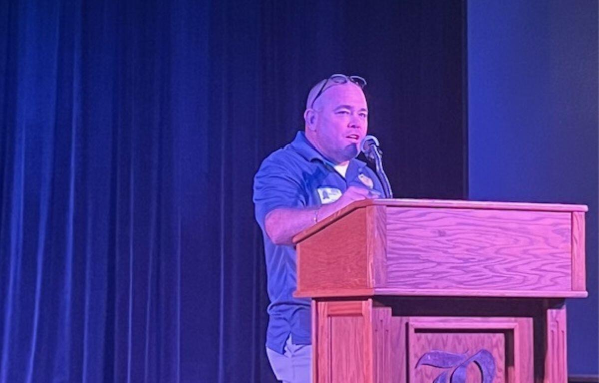 A man in a blue shirt standing behind a podium with a carved wooden insignia on the front speaking into a microphone with a blue curtain in background.