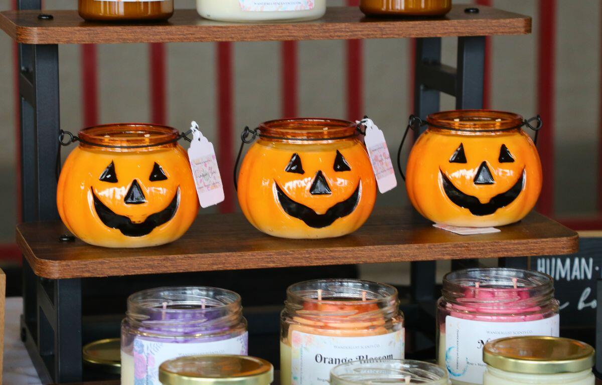 Three pumpkin candles, scented to pumpkin spice are lined up on a wooden shelf with other scents surrounding them.