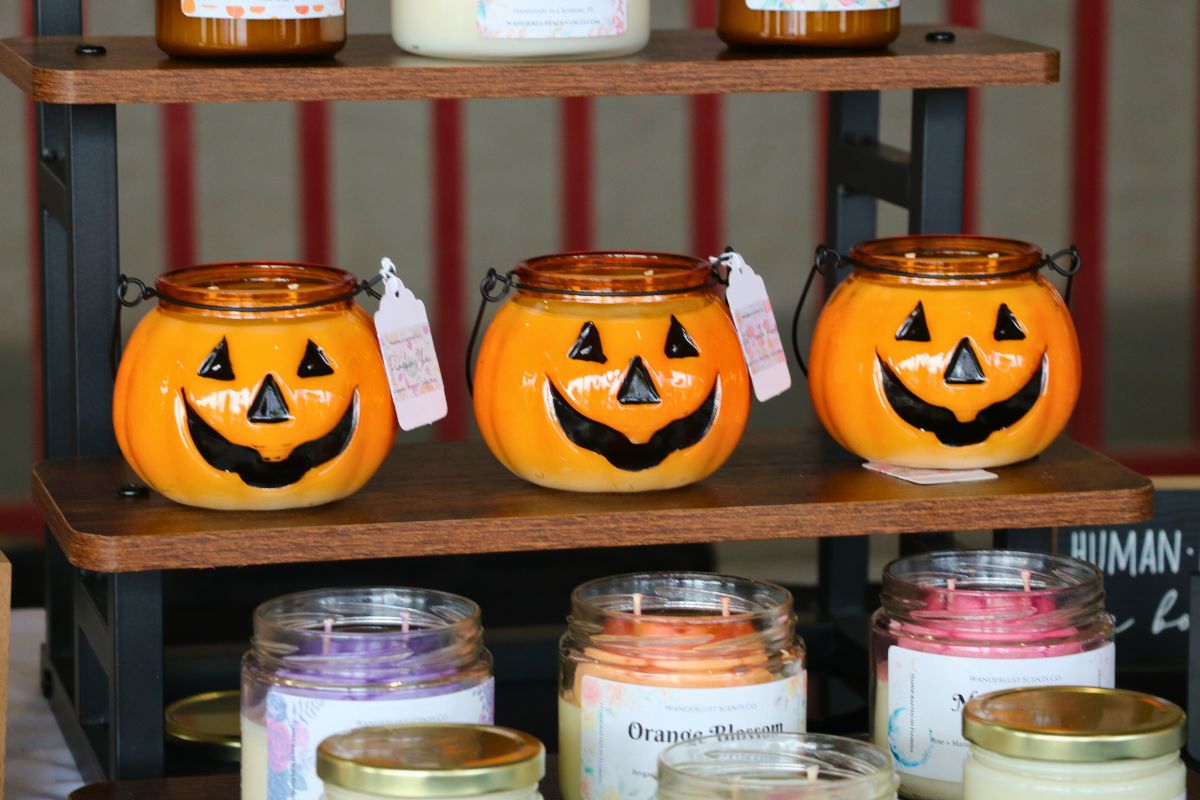 Three pumpkin candles, scented to pumpkin spice are lined up on a wooden shelf with other scents surrounding them.