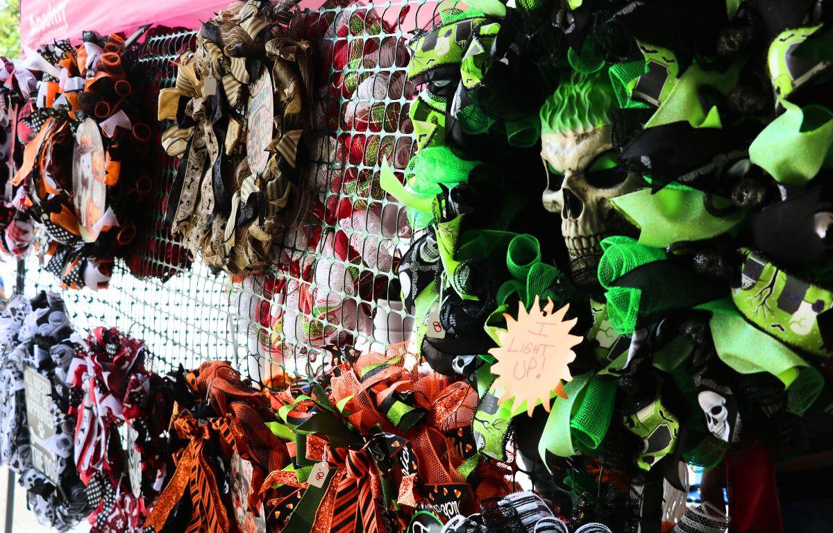 A green and black wreath with a skull in the center is hung on a rack with a sign that reads, "I Light Up!" To the right, more wreaths are hung in a colorful display of fall themes and colors.