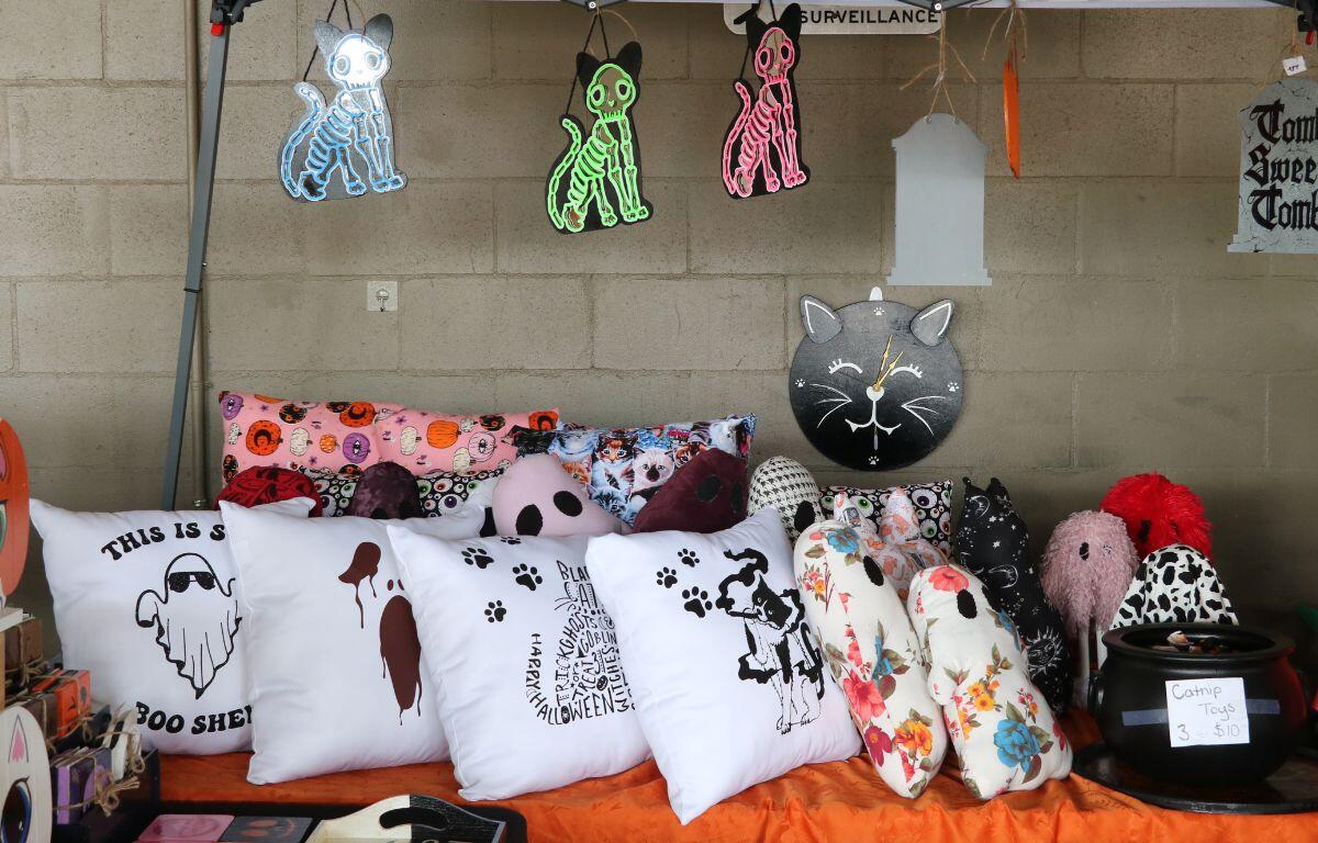 Skeletal cat lights in varying colors and tomb stones are hung from a canopy above a table. The table is covered in an orange tablecloth. On top of the table are Halloween-themed pillows and a cauldron that reads, "Catnip Toys."