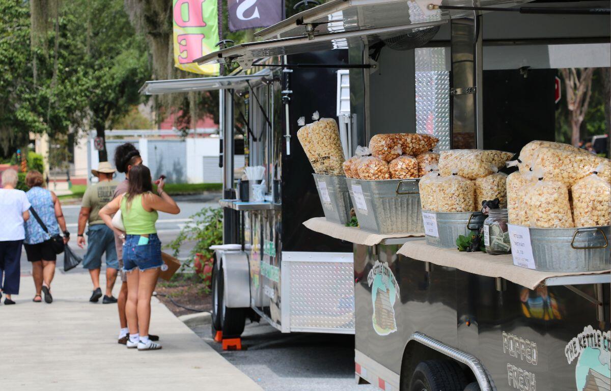 Kettle corn bags sit in buckets on the edge of a counter at a food truck parked on the curb of a sidewalk. In the background, two people are ordering from a lemonade stand while others walk out toward a street.