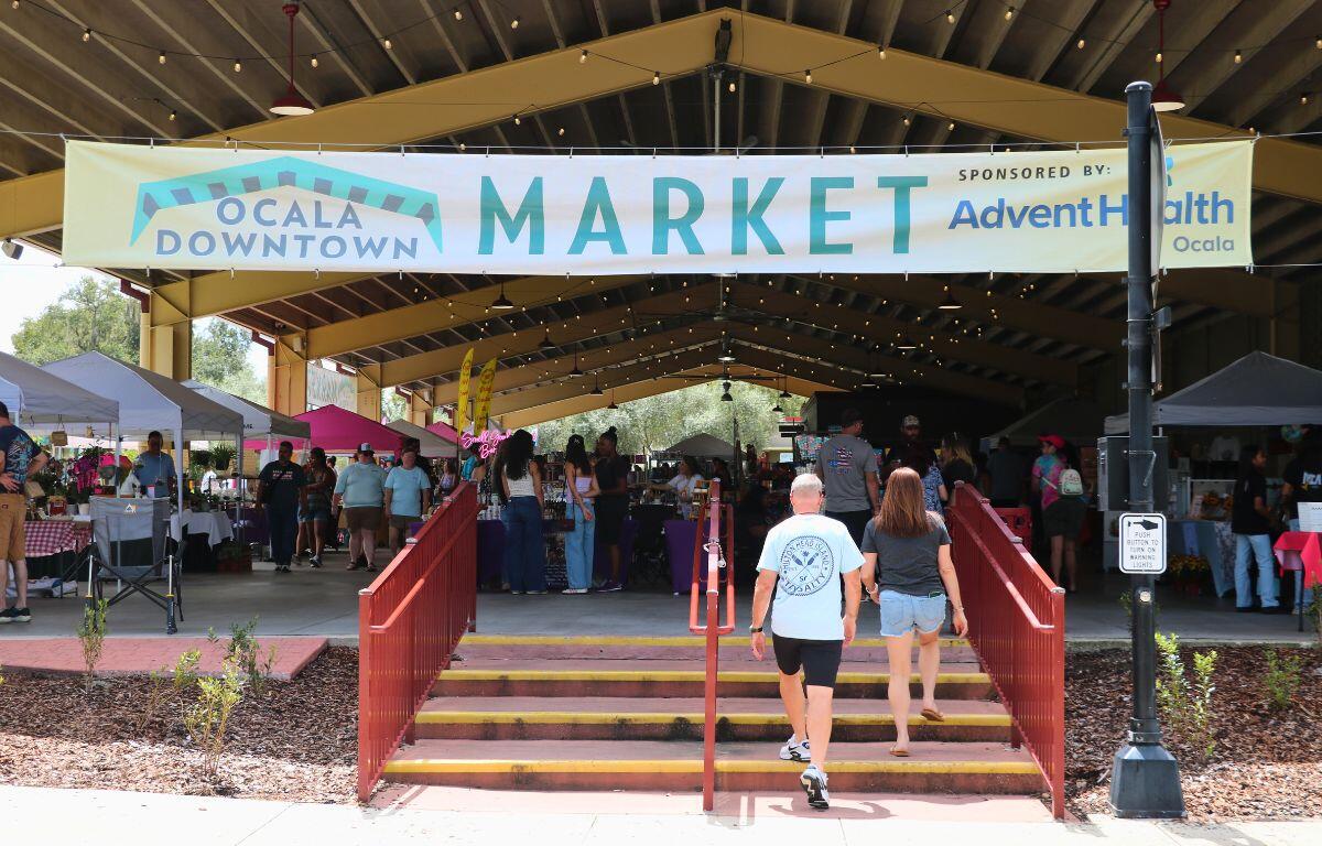 A large white banner is hung across an open pavilion reading, "Ocala Downtown Market Sponsored by: Advent Health Ocala." Under the pavilion are vendor lining the edges and center while guests browse at the multitude of booths. Two people can be seen walking toward the market under the banner.