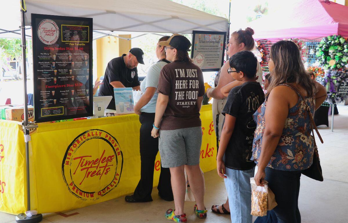 People line up at a booth with a white canopy on top, signs reading the drinks that are offered and a table with yellow signs hanging around it reading, "Timeless Treats."