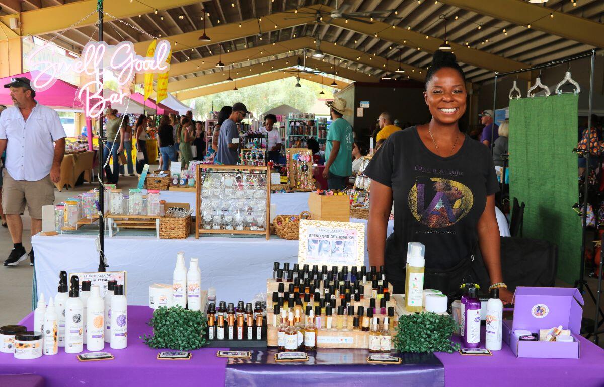 A woman stands behind a table with a purple tablecloth. On the table is hair products and essential oils lined up with a sign behind it reading, "Smells good Bar." In the background are various other booths selling items ranging from jewelry to cups.
