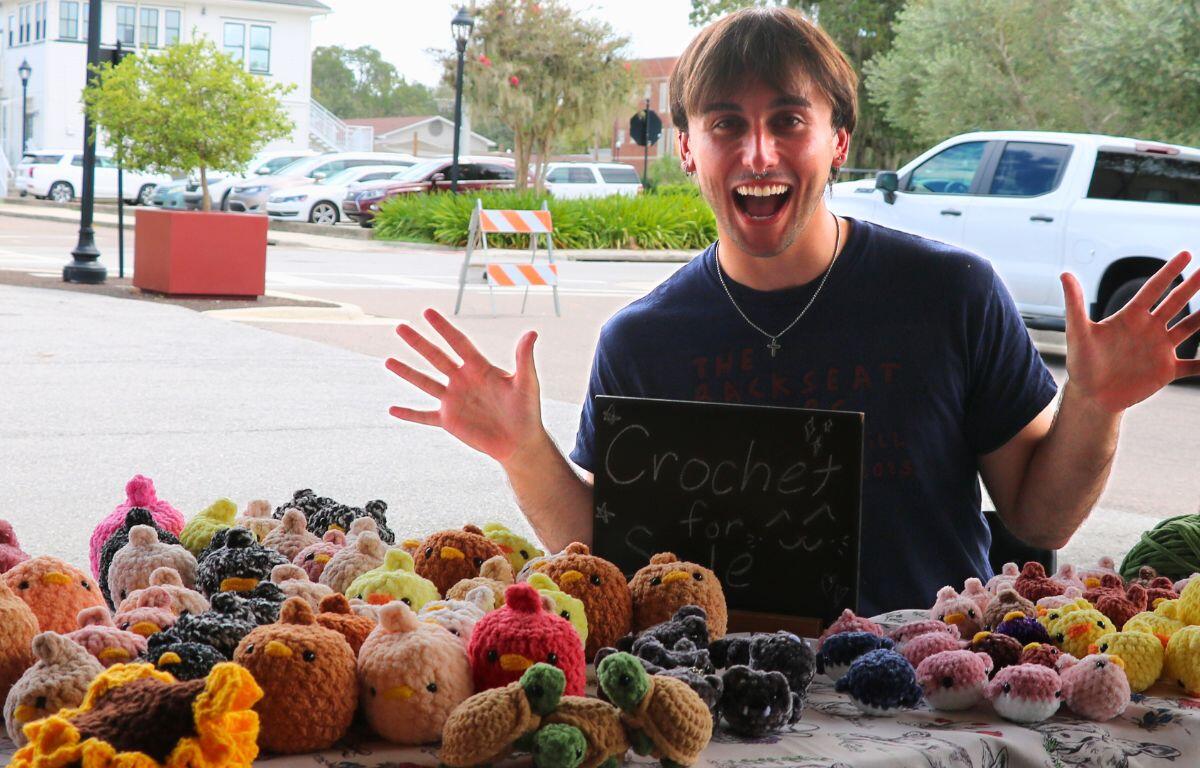 A young man sits behind a table with multiple crochet animals and sunflower coasters. In the background is a white truck, orange and yellow traffic barricade, a potter with a small tree and a parking lot.