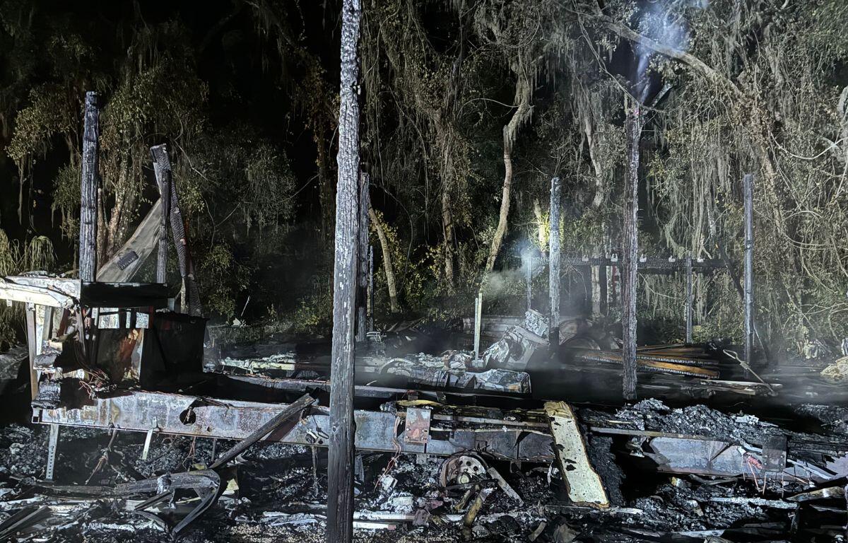 Tree line in the background, smoldering gray remains, of a barn, with a pole and debris in the foreground.
