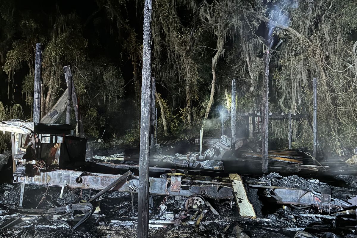Tree line in the background, smoldering gray remains, of a barn, with a pole and debris in the foreground.