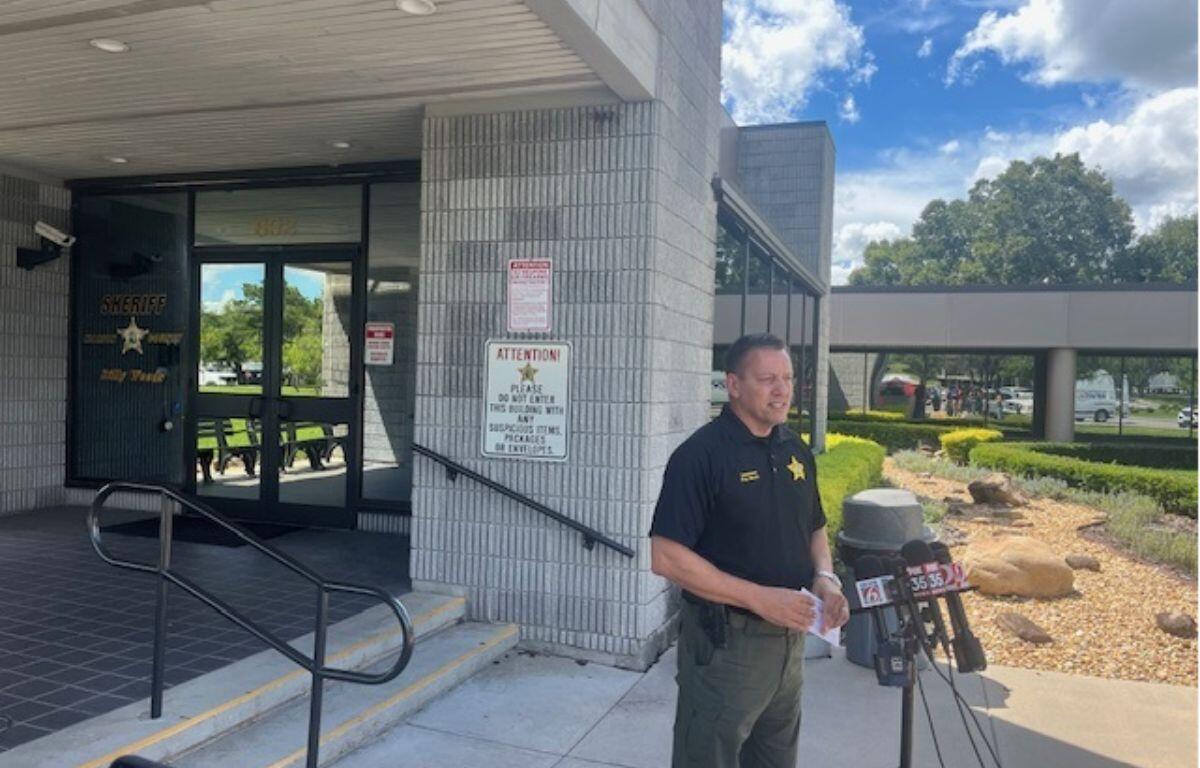 Glass doors with an insignia, a concrete wall, with a sign on the left, the subject in the in middle standing before a battery of microphones, with a trash receptacle, and curb appeal landscaping to the right. On the far-right vehicles in a parking lot.