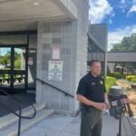Glass doors with an insignia, a concrete wall, with a sign on the left, the subject in the in middle standing before a battery of microphones, with a trash receptacle, and curb appeal landscaping to the right. On the far-right vehicles in a parking lot.
