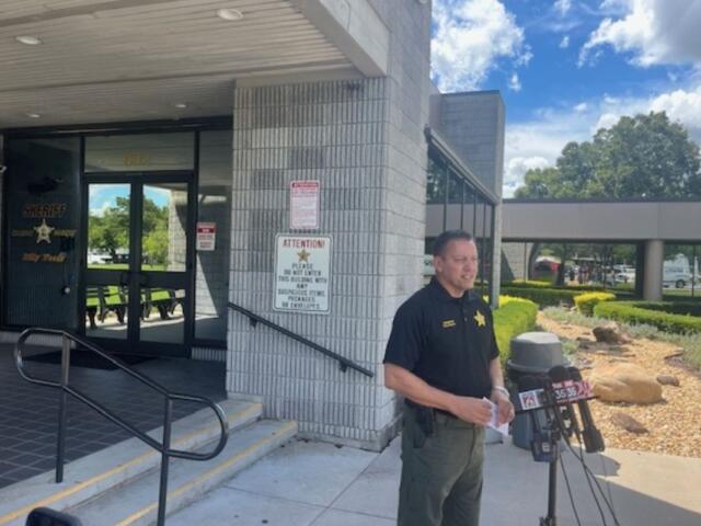 Glass doors with an insignia, a concrete wall, with a sign on the left, the subject in the in middle standing before a battery of microphones, with a trash receptacle, and curb appeal landscaping to the right. On the far-right vehicles in a parking lot.