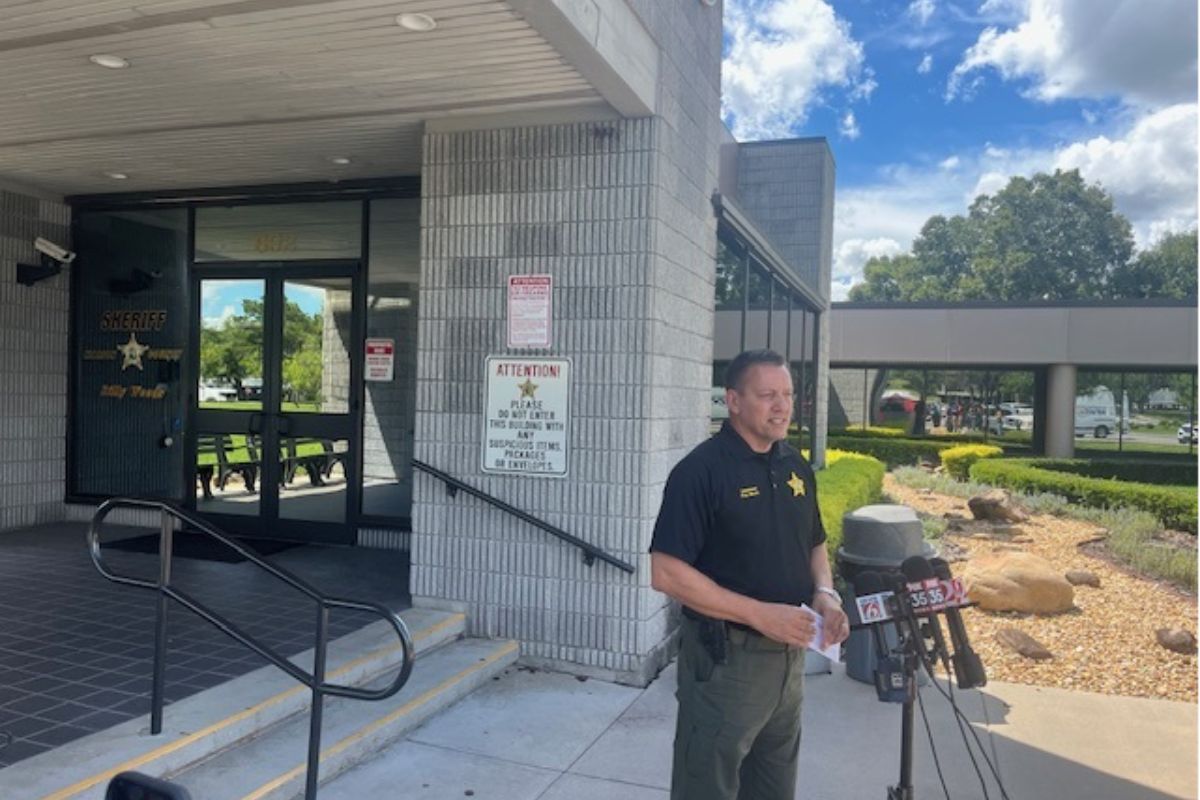 Glass doors with an insignia, a concrete wall, with a sign on the left, the subject in the in middle standing before a battery of microphones, with a trash receptacle, and curb appeal landscaping to the right. On the far-right vehicles in a parking lot.