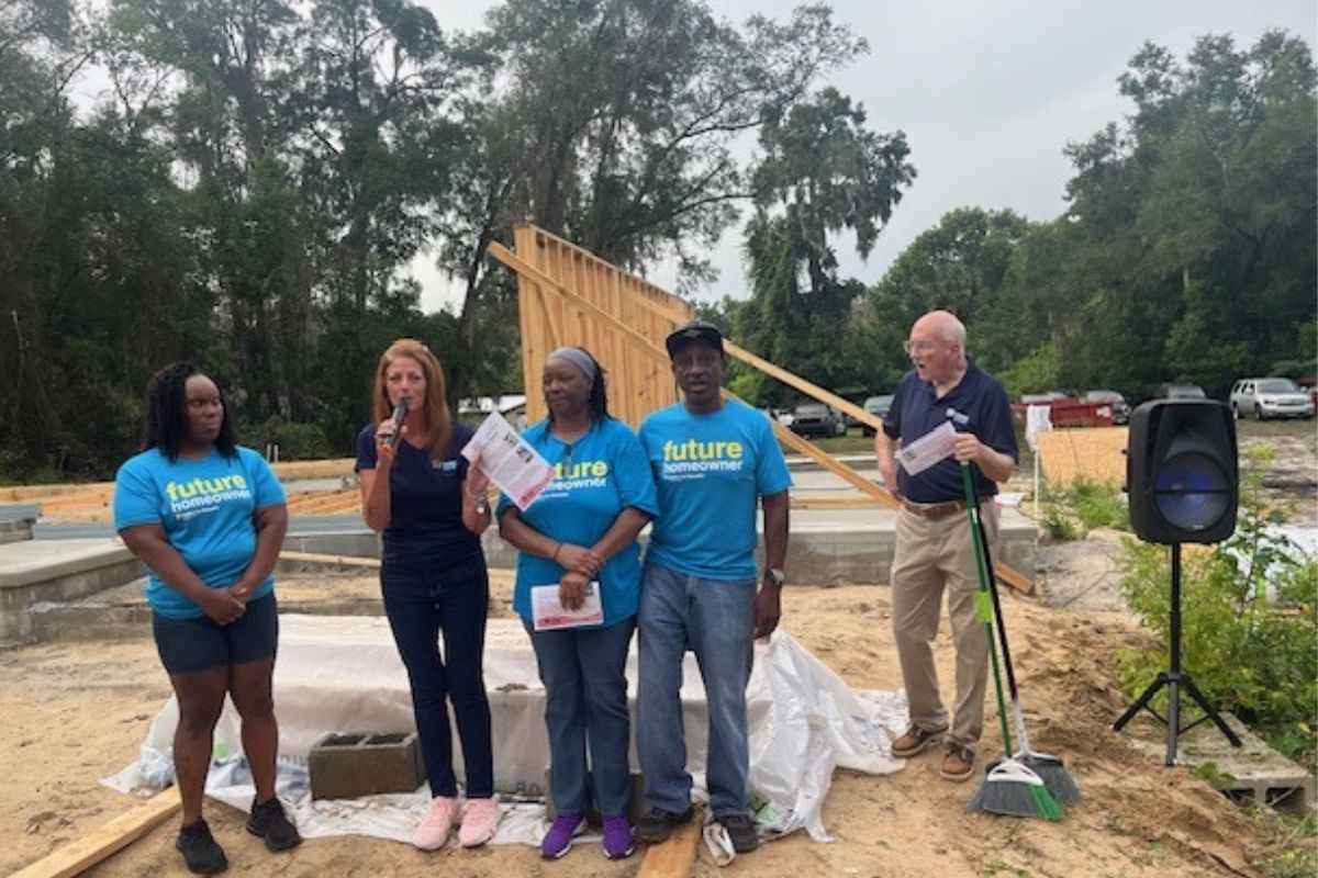 A woman in a blue t-shirt, next to a woman holding a microphone, with a man and a woman to her right, with a man to their right holding a broom, with a wood frame in the background, with a tree line in the far background. 