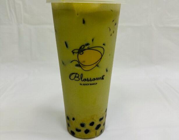 A green drink in a plastic cup sits on a table with a white tarp and white background. The cup has a graphic of fruit with "Blossoms Juicy Bar" written under it. Black tapioca pearls lay at the bottom of the cup while ice floats toward the top.
