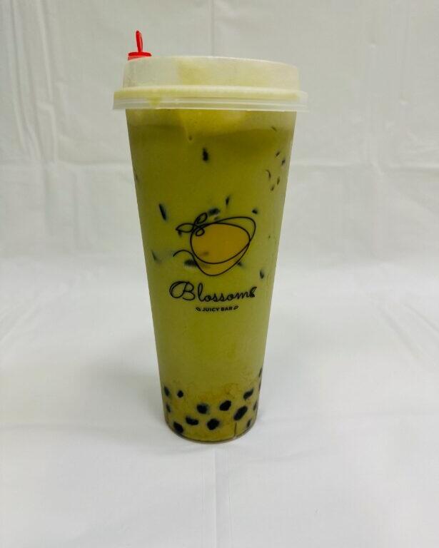 A green drink in a plastic cup sits on a table with a white tarp and white background. The cup has a graphic of fruit with "Blossoms Juicy Bar" written under it. Black tapioca pearls lay at the bottom of the cup while ice floats toward the top.