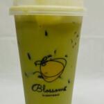A green drink in a plastic cup sits on a table with a white tarp and white background. The cup has a graphic of fruit with "Blossoms Juicy Bar" written under it. Black tapioca pearls lay at the bottom of the cup while ice floats toward the top.