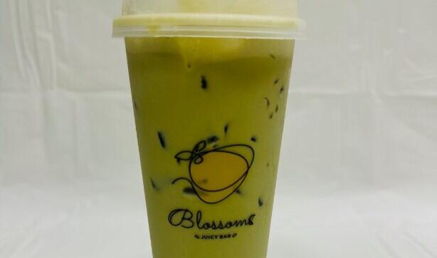 A green drink in a plastic cup sits on a table with a white tarp and white background. The cup has a graphic of fruit with "Blossoms Juicy Bar" written under it. Black tapioca pearls lay at the bottom of the cup while ice floats toward the top.