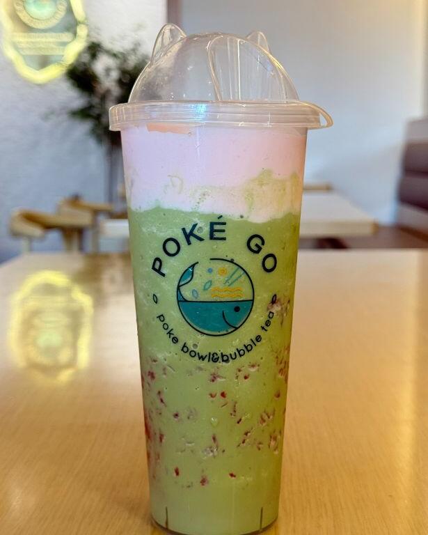A plastic cup sits on a wooden table with a cafe setting in the background. On the sup reads, "Poke Go. Poke bowl & bubble tea." Inside the cup is a green drink with pink foam floating on the top and strawberry bubbles floating in the middle and bottom.