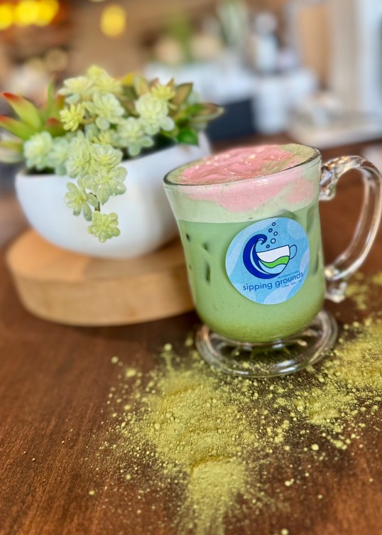 A glass cup with a blue sticker on the front has a graphic of a cup of tea with "Sipping Grounds" written under it. On the table in front of it is matcha powder. Behind it on the table is a flower in a planter on a wooden lift with a cafe in the background. Inside the glass is a green drink with ice and a light pink addition to the drink at the top.