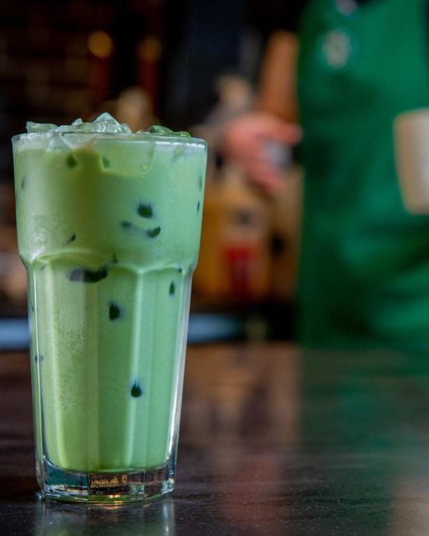 A clear glass cup stands on a dark table with a green drink on the inside and ice floating toward the top. In the background is a barista in a green apron holding another cop.