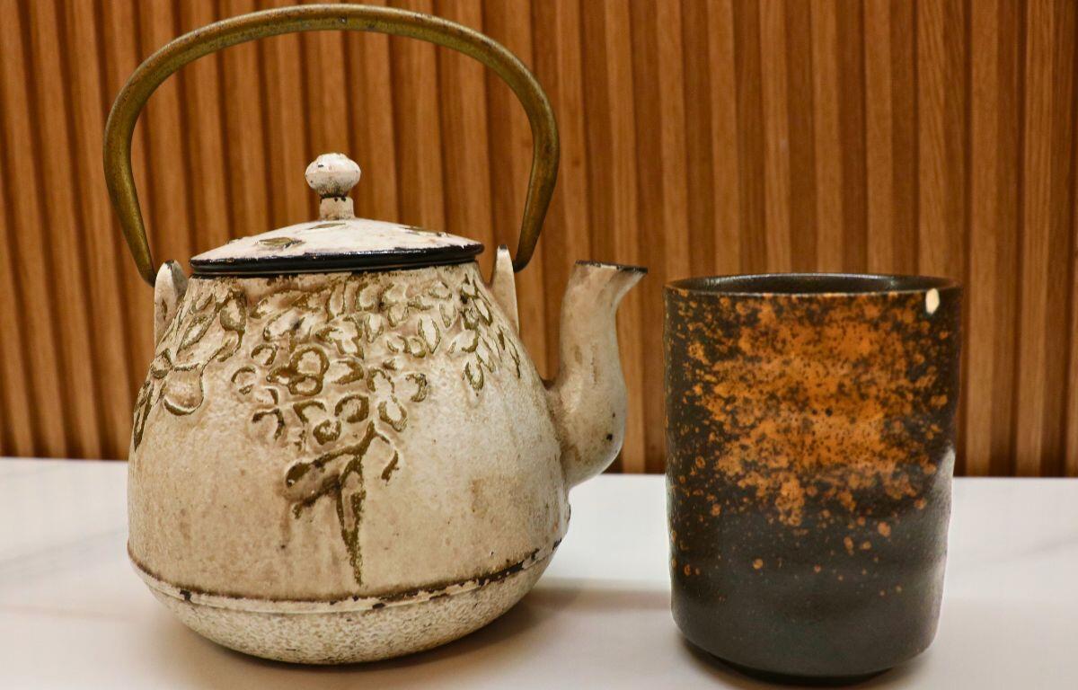 A white teapot with gray designs and a bronze on the left and a black teacup on the right with bronze accents. In the background is a brown wood wall and a white table.