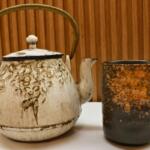 A white teapot with gray designs and a bronze on the left and a black teacup on the right with bronze accents. In the background is a brown wood wall and a white table.