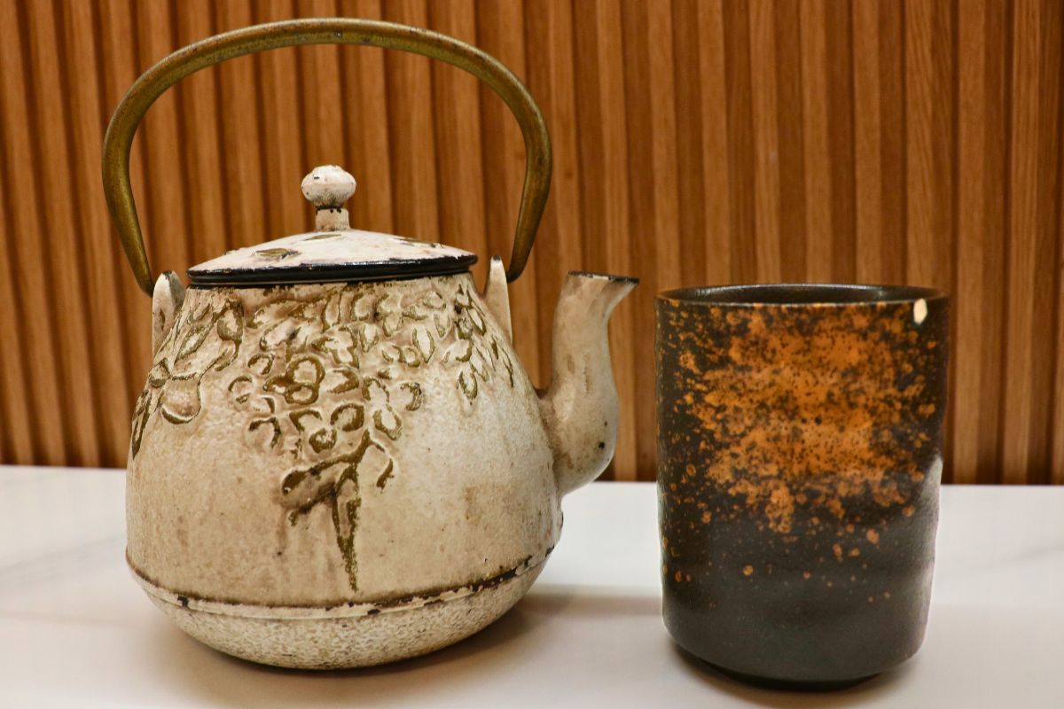 A white teapot with gray designs and a bronze on the left and a black teacup on the right with bronze accents. In the background is a brown wood wall and a white table.