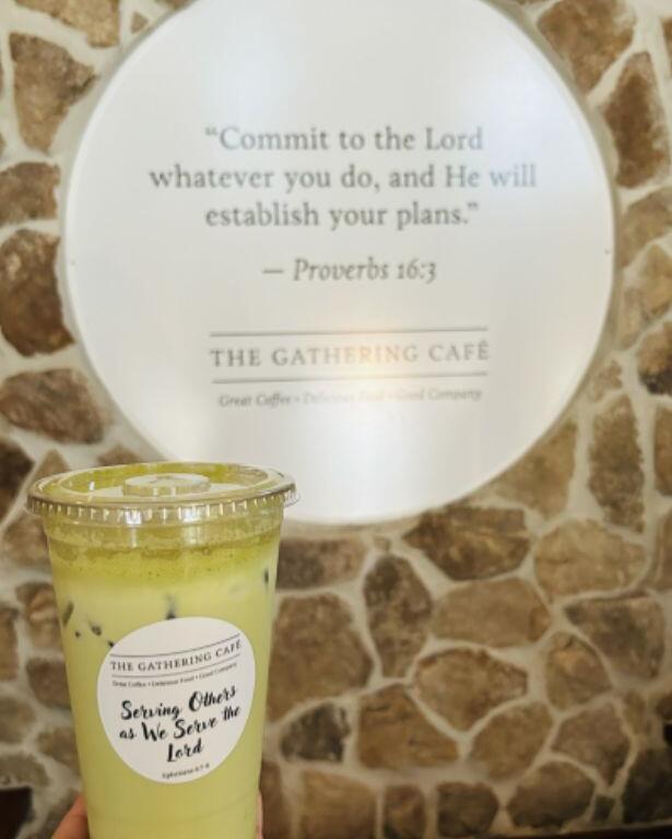 A clear plastic cup holds a green drink with ice floating in it. On the front of the cup is a stick that reads, "The Gathering Cafe. Serving Others as We Serve the Lord." In the background is a stone wall with a large white sign behind it reading Proverbs 16:3 and "The Gathering Cafe."