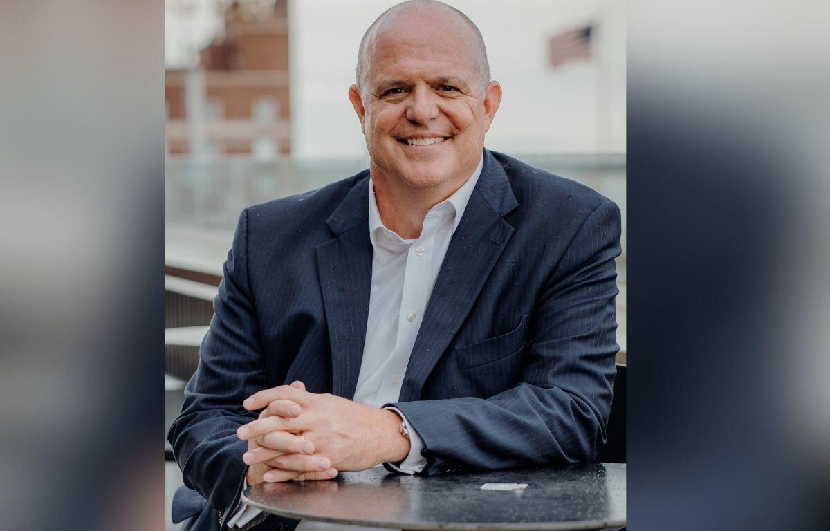 Well-dressed gentleman with his hands on a table, smiling, with an out of focus United States of America over his right shoulder and an out of focus building to his left.
