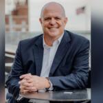 Well-dressed gentleman with his hands on a table, smiling, with an out of focus United States of America over his right shoulder and an out of focus building to his left.