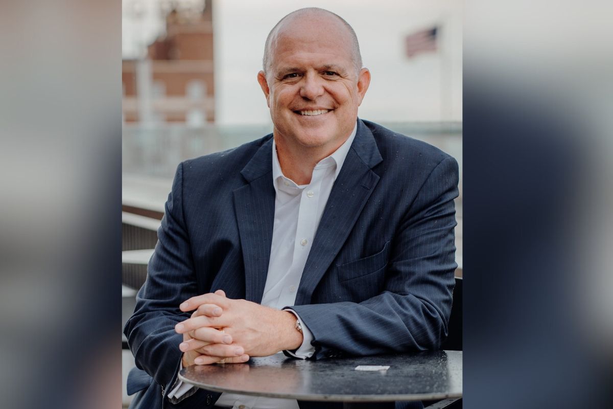 Well-dressed gentleman with his hands on a table, smiling, with an out of focus United States of America over his right shoulder and an out of focus building to his left.