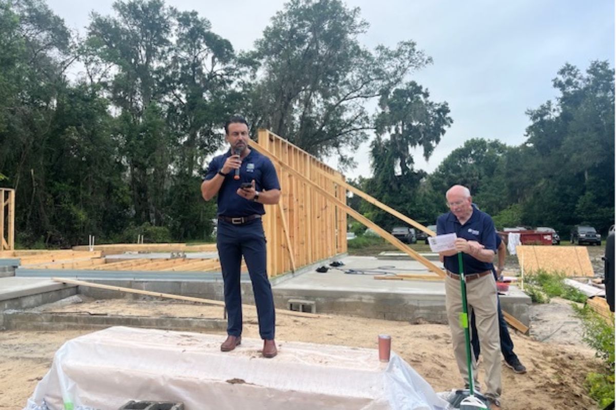 In the background, a skyline with a tree, a frame on top of a concrete foundation, and a treeline, in the foreground, a man stands on a raised platform holding a microphone and phone, and man with a broom to the right.