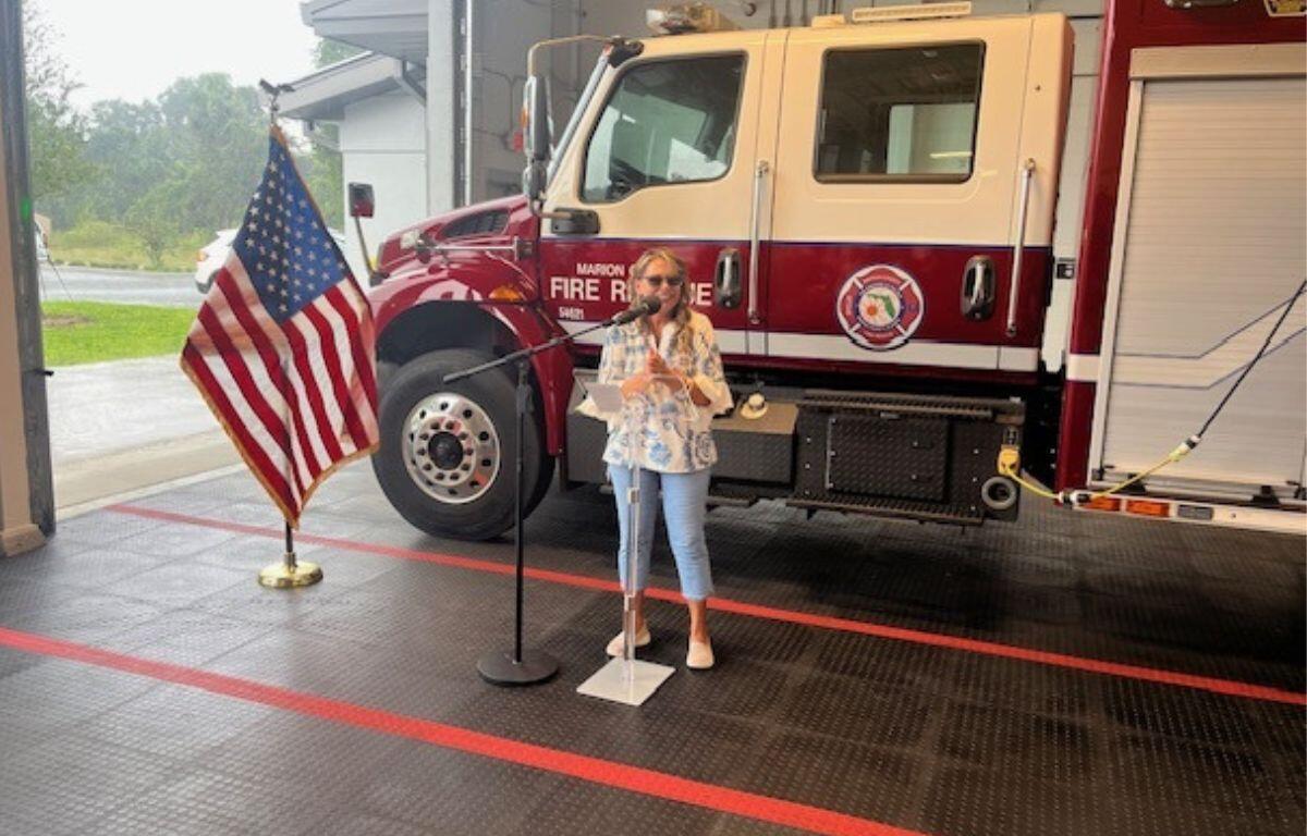 To the left Flag of the United States of America. In the background a Marion County Fire and Rescue Truck. A concrete floor with orange stripes in the foreground. Subject standing behind a microphone.