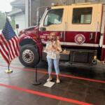 To the left Flag of the United States of America. In the background a Marion County Fire and Rescue Truck. A concrete floor with orange stripes in the foreground. Subject standing behind a microphone.