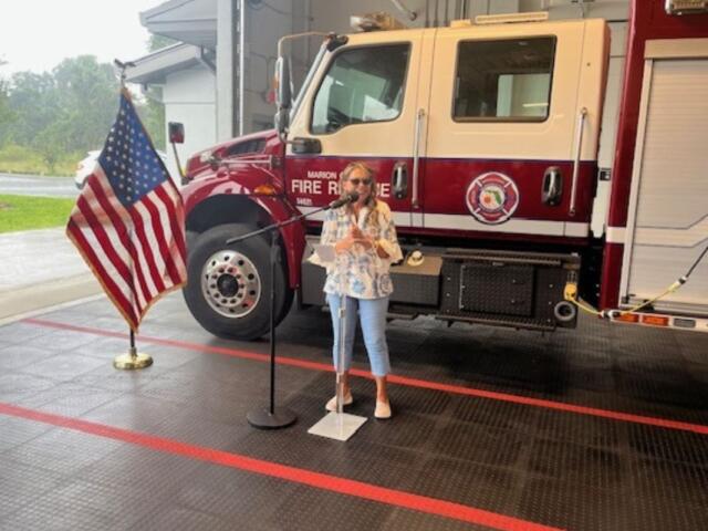 To the left Flag of the United States of America. In the background a Marion County Fire and Rescue Truck. A concrete floor with orange stripes in the foreground. Subject standing behind a microphone.