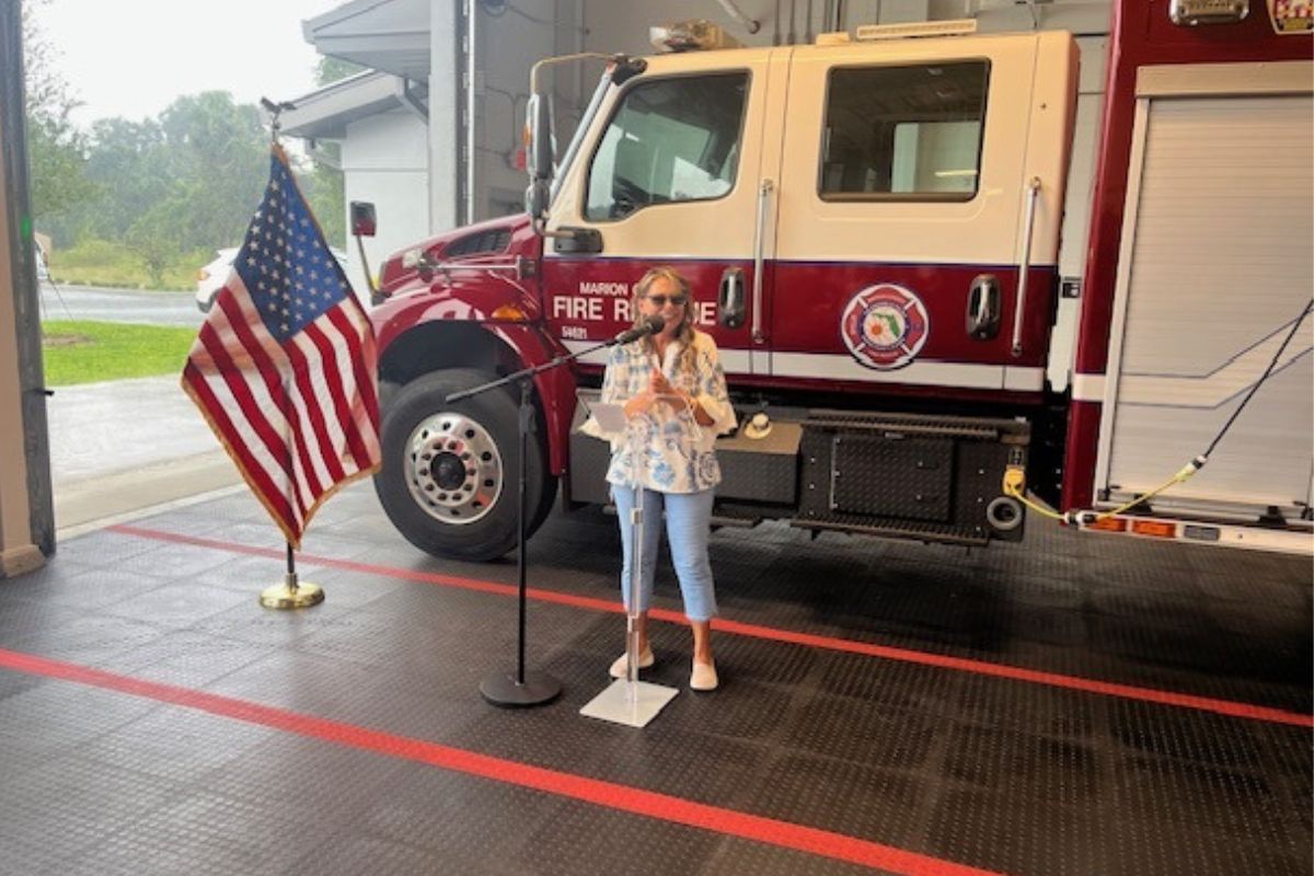 To the left Flag of the United States of America. In the background a Marion County Fire and Rescue Truck. A concrete floor with orange stripes in the foreground. Subject standing behind a microphone.