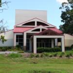 Front view of modern building with a beige exterior, red metal roof, large windows, and symmetrical entrance surrounded by landscaped greenery.