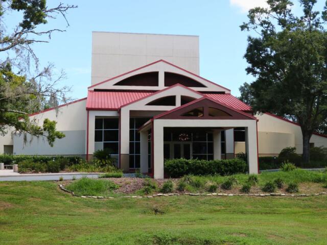 Front view of modern building with a beige exterior, red metal roof, large windows, and symmetrical entrance surrounded by landscaped greenery.