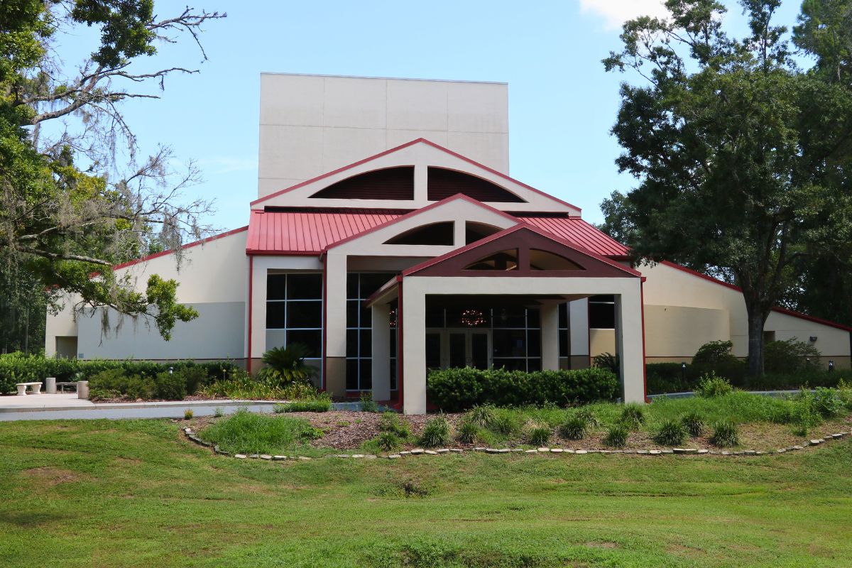 Front view of modern building with a beige exterior, red metal roof, large windows, and symmetrical entrance surrounded by landscaped greenery.