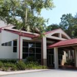 Front view of modern building with a beige exterior, red metal roof and large windows with trees and greenery surrounding it.