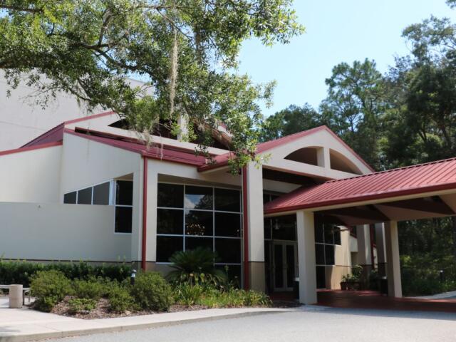 Front view of modern building with a beige exterior, red metal roof and large windows with trees and greenery surrounding it.