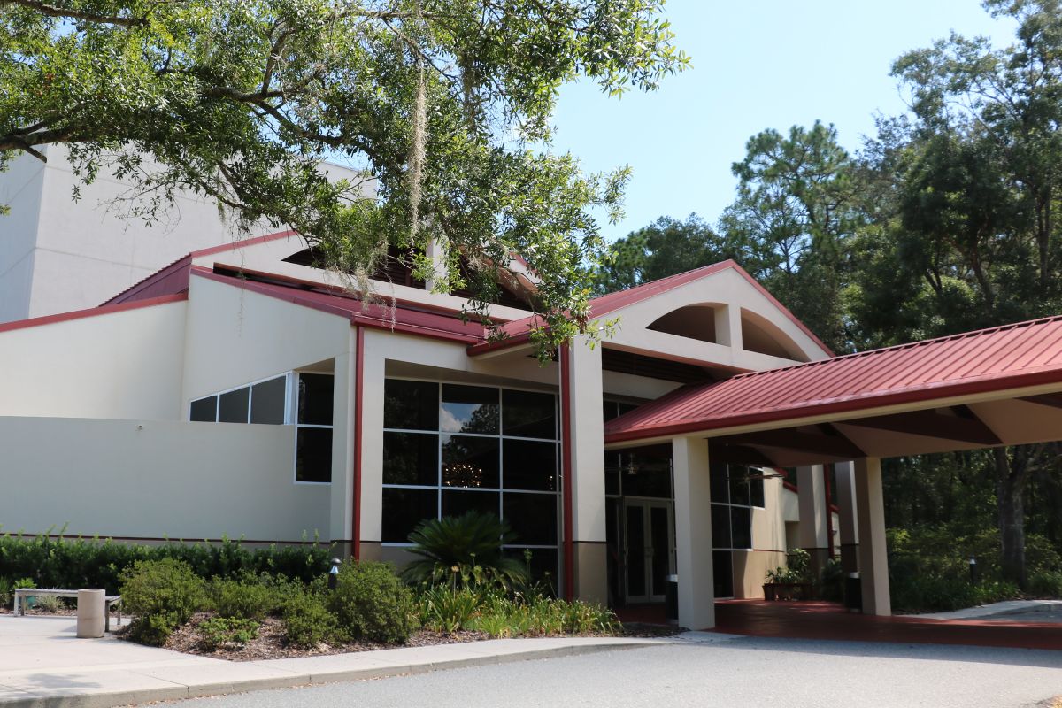Front view of modern building with a beige exterior, red metal roof and large windows with trees and greenery surrounding it.