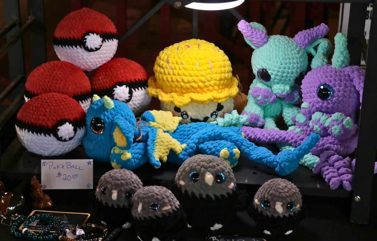 Handcrafted crochet plushies sit on a black shelf with a light shining to showcase their features. These items include Poke Balls, a mushroom, dragons and birds. Toward the bottom jewelry can be seen on a table with a black tablecloth.