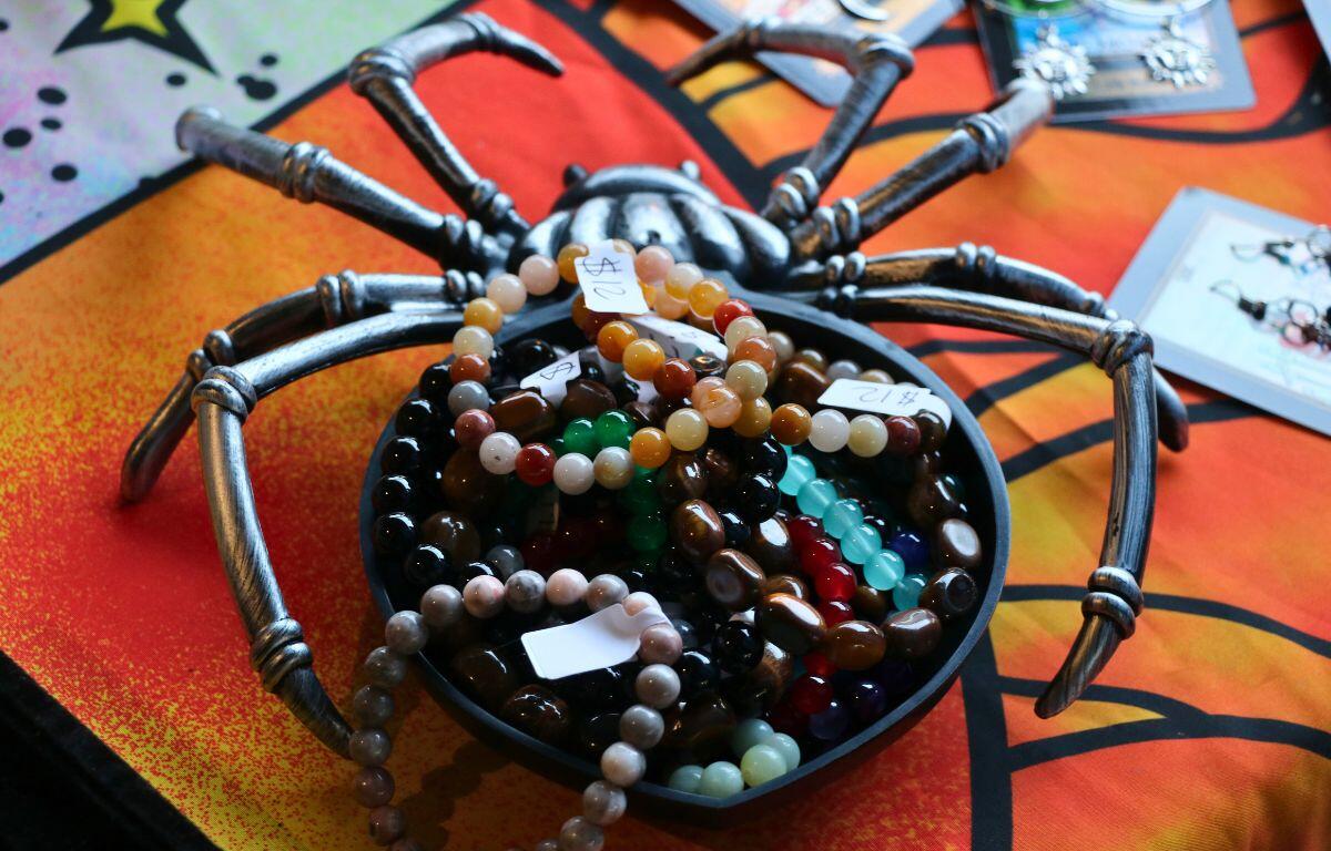 A gray spider-shaped bowl is filled to the brim with handcrafted bracelets on a table with an orange-patterned tablecloth. In the background, earrings can be seen laid out on the table.