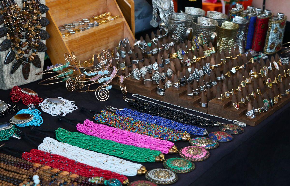 A colorful display of handmade jewelry at a market stall, featuring an assortment of rings, bracelets, necklaces, and decorative accessories. The setup includes wooden stands holding silver and gold rings, beaded necklaces in vibrant colors, large decorative pendants, and ornate metal cuffs and flasks. The background shows a variety of textures and materials, suggesting a bohemian or artisanal theme.