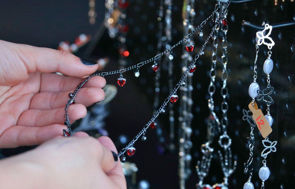 Two hands hold a necklace with red hearts strung around it. In front of the necklace is another necklace with butterflies and a white jewel str4ung around it with a price tag that reads, "12". Blurred in the background are a variety of other necklaces with different designs.