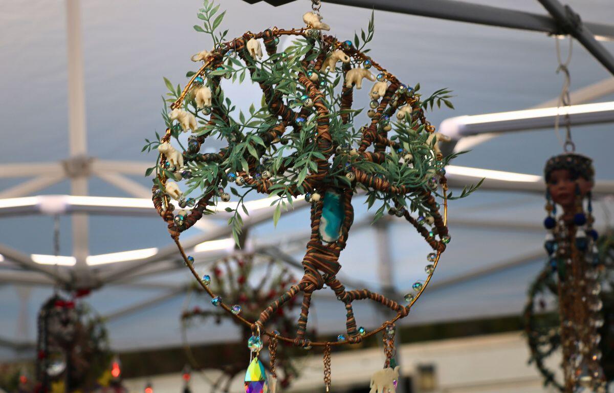 A circular dream catcher hosts a tree in the center with 4 pieces meant to represent roots coming up to a single section at the center meant to represent the center of a tree before reaching up into multiple sections representing branches with leaves and handcrafted animal figures. In the background, more dream catchers can be seen hanging from a tent with lights strewn on the support beams.