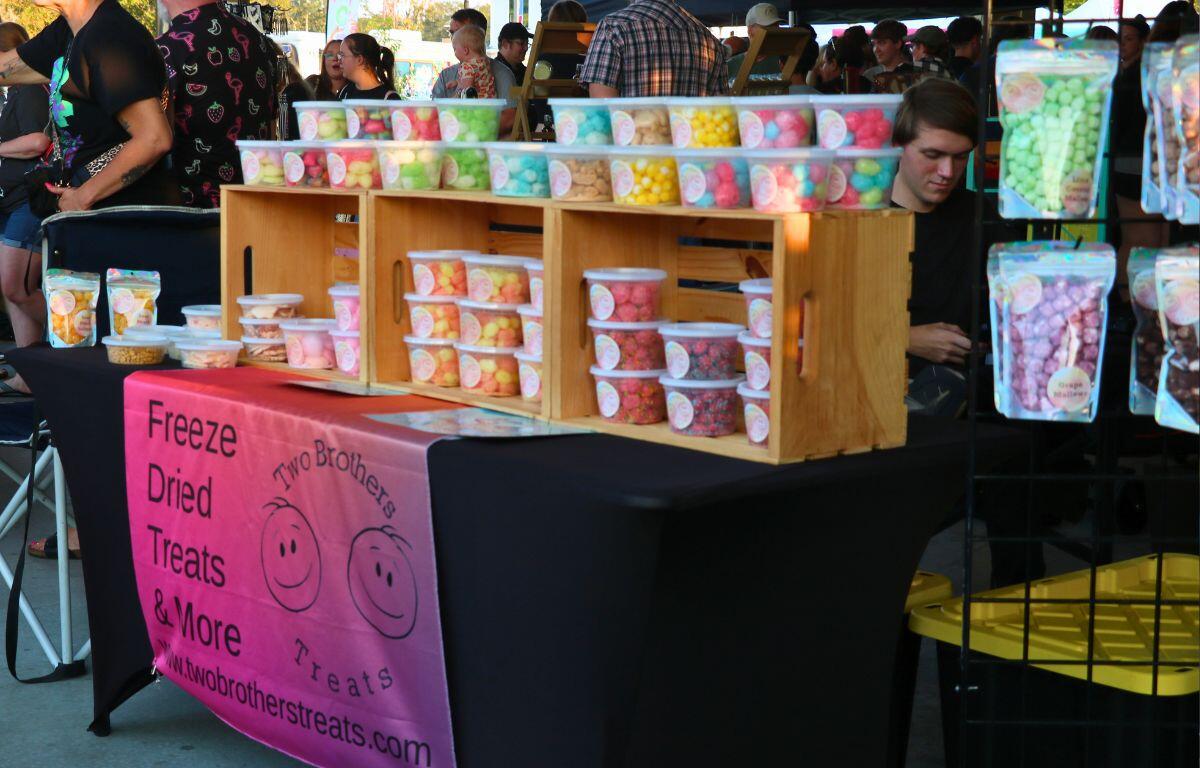A variety of freeze dried treats sit on a table with a black tablecloth and hang from a black rack. Guests shop through a market setting in the background.